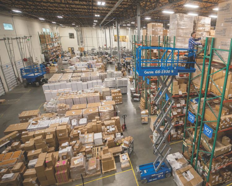 A Genie scissor lift extended to the top rack inside a warehouse, with another scissor lift in the background.