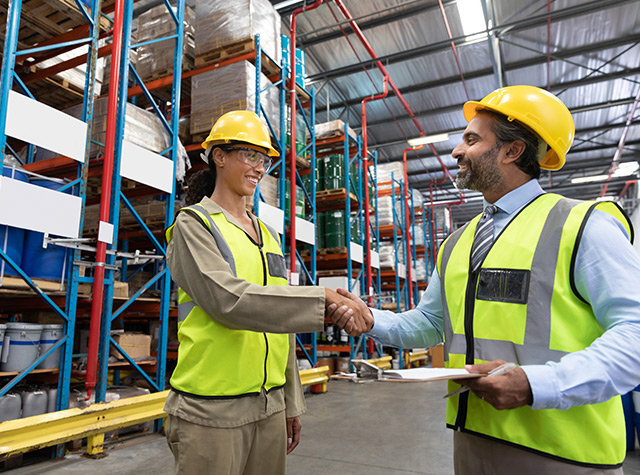 Two individuals shaking hands inside a warehouse standing next to pallet racking.