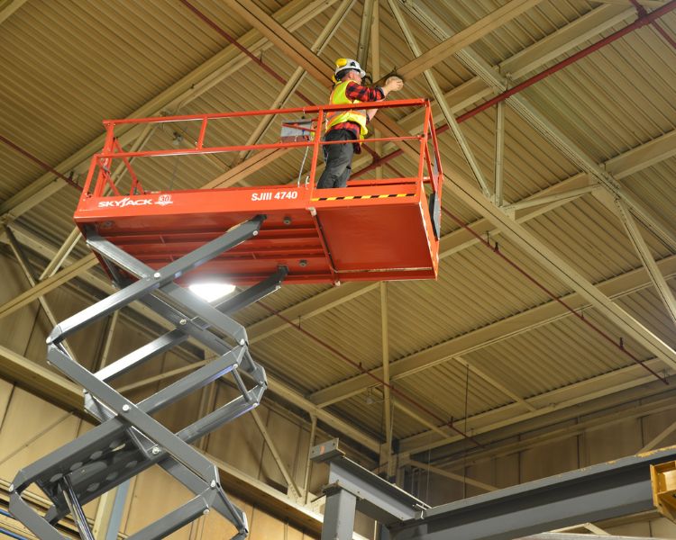 A Skyjack scissor lift extended to the ceiling with an operator on the platform.
